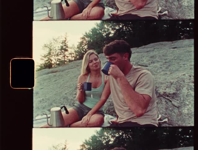 A woman and a man sit on a rocky surface outdoors drinking from mugs together near a coffee pot