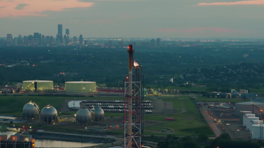 An aerial view of a refinery flare stack with a city skyline at dusk