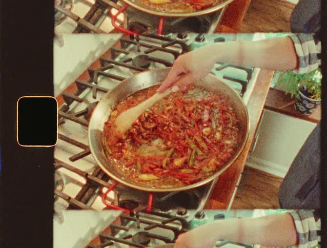 A person stirring pan of vegetables as sauce simmers on a stovetop filmed with a grainy vintage effect