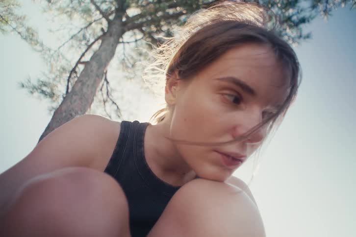 In a lowangle closeup a woman under a tree looks down with a pensive expression her face briefly washed out by a sun flare before she closes her eyes