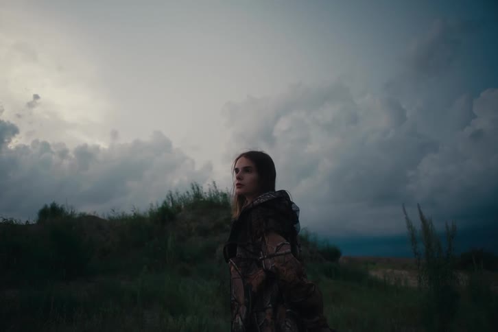 A young woman in a camouflage jacket stands in a grassy field at dusk gazing toward the cloudy darkening sky