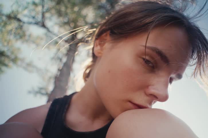 A young woman with loose hair leans her face against her knee in soft daylight her expression appearing thoughtful and subdued