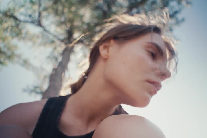 A woman sits under a tree in sunlight looking pensive