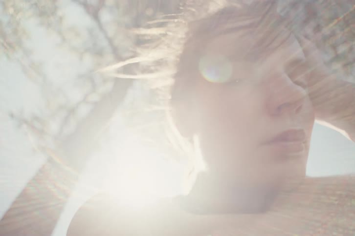 A young woman sits outdoors under a tree with the sun reflecting on her at midday