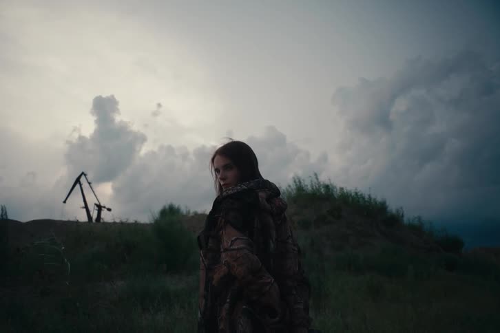 Woman in a camouflage coat walks through an open field at dusk looking back