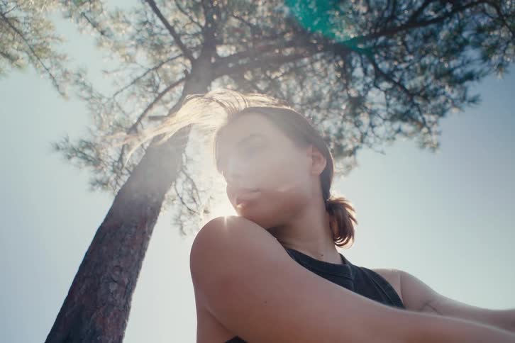 En young woman looks to the side with strands of hair blowing in the wind near a sunlit tree