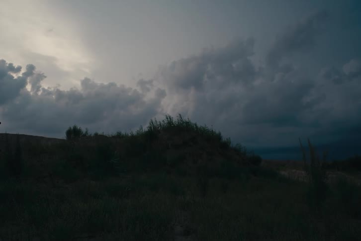 A grassy hill is silhouetted against a dramatic cloudy sky at dusk