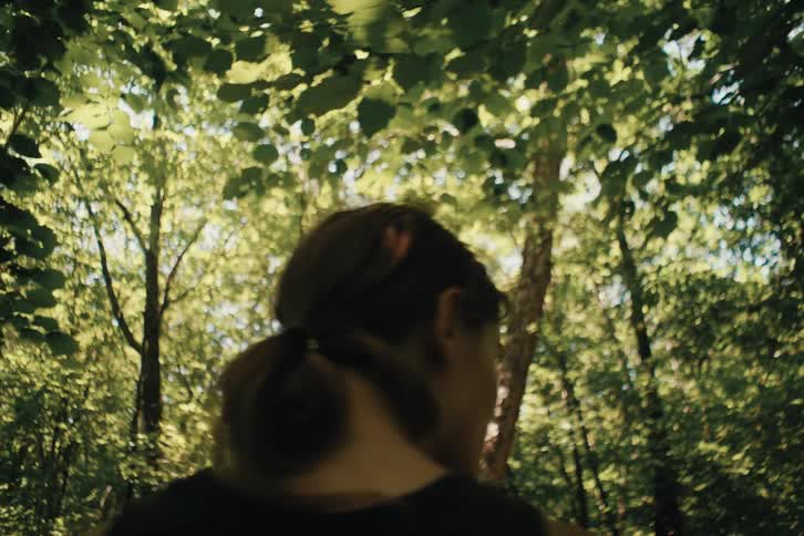 The video shows a person with a top knot hairstyle wearing a dark tank top walking through a forest with dense foliage sunlight filters through the canopy creating a dappled bright effect on the leaves