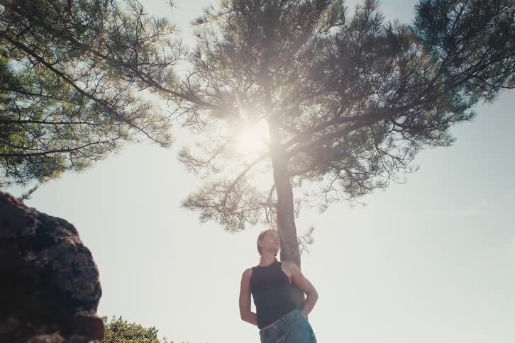 A video shows a lowangle shot of a woman with her back to a tree looking up the sun is shining brightly from behind the tree creating a strong lens flare the woman is wearing a dark tank top and jeans