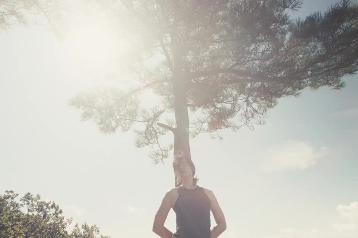 A young woman is standing outdoors looking directly at the camera with a calm expression as bright sunlight filters through a tree above her