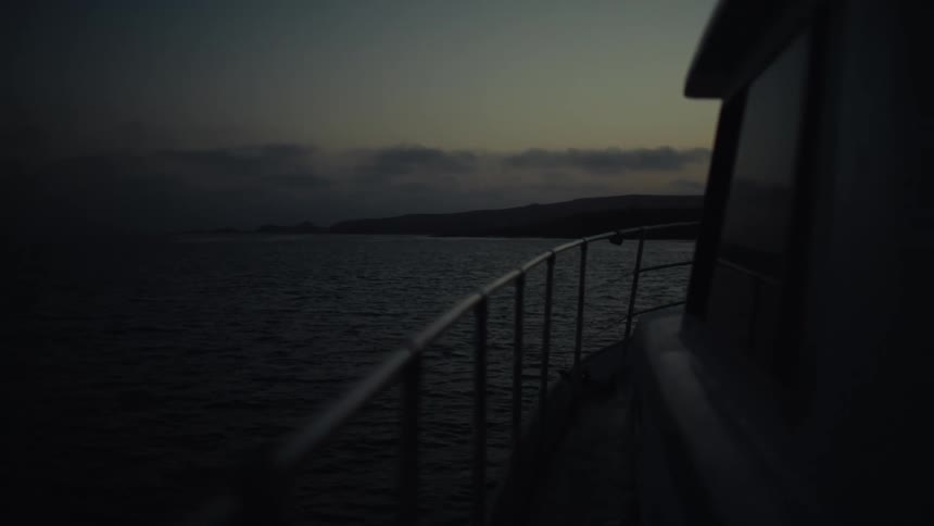 A boat glides across dark waters at dusk with the railing and cabin visible in the fading light against distant hills