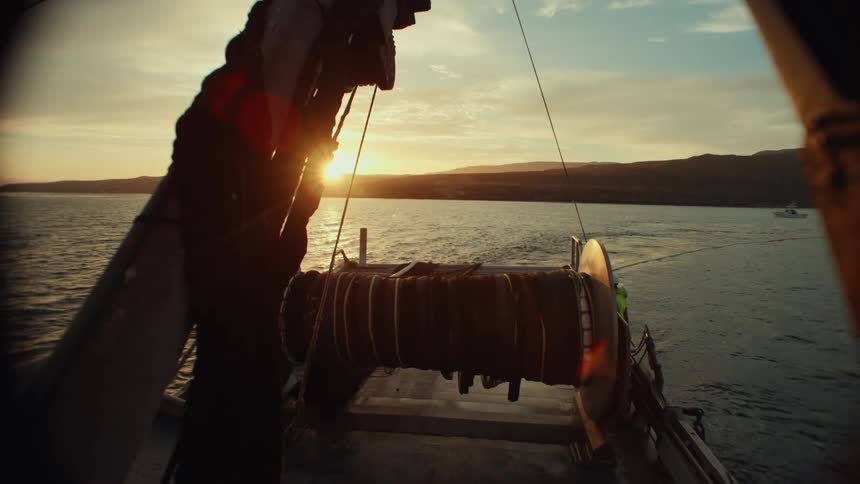 A fishing boat heads across the water at sunset with large nets wound on a spool and distant hills silhouetted against the sky