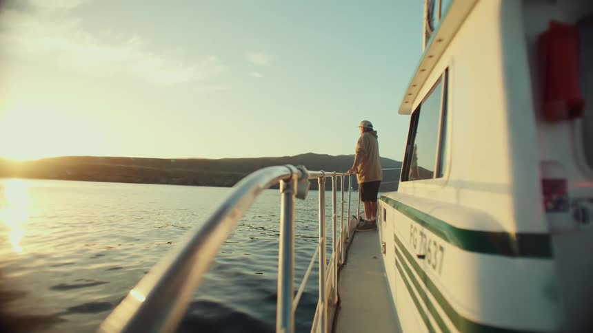 Person standing near the rail of a boat stares towards the sun sitting on the horizon