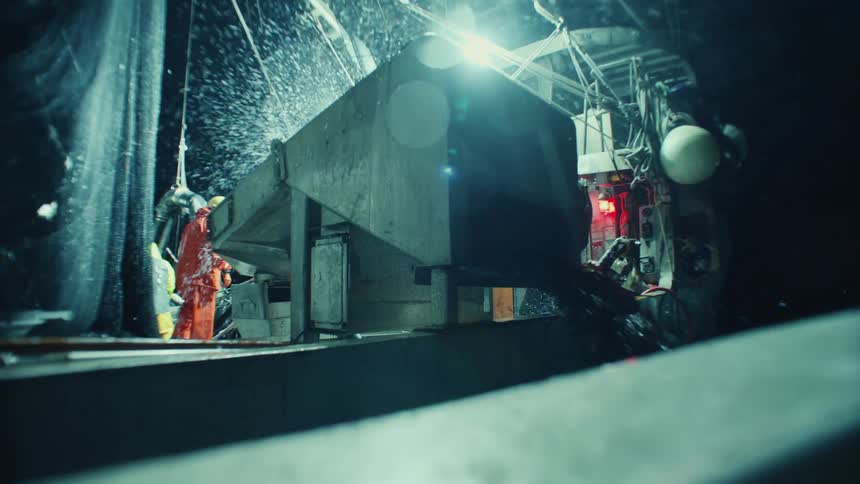 A factory worker wearing a raincoat and hard hat stands operating a machine that is spilling liquid at night