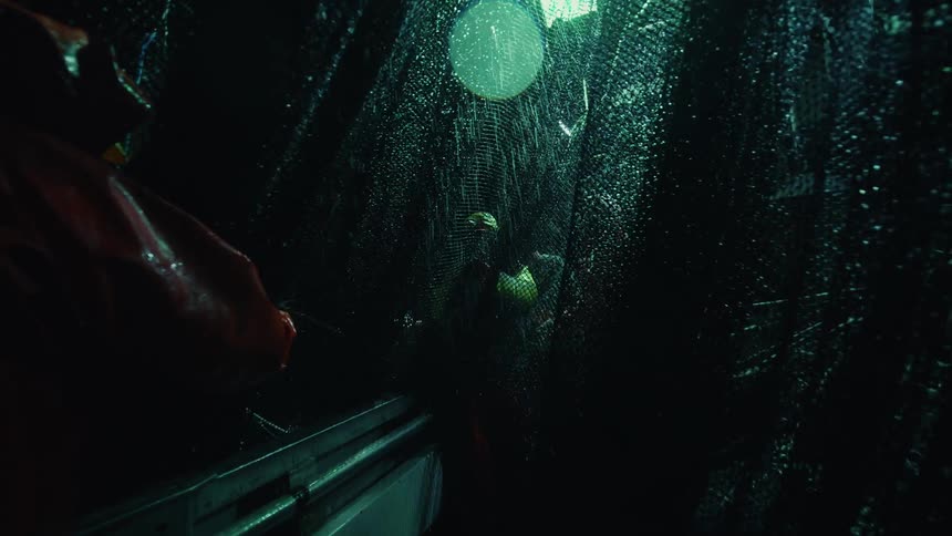 At night during a storm commercial fishermen in heavy rain gear work on the wet deck of a fishing boat next to a large glistening net