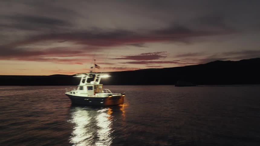 A boat illuminated by bright lights drifts on dark water at sunset casting a long reflection beneath a vivid sky streaked with clouds