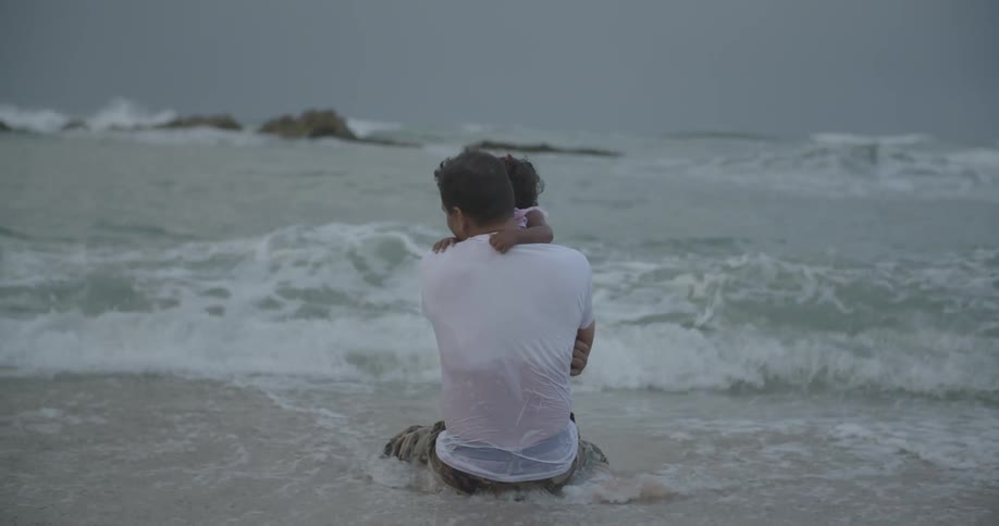 A man and child sit in the water of a beach