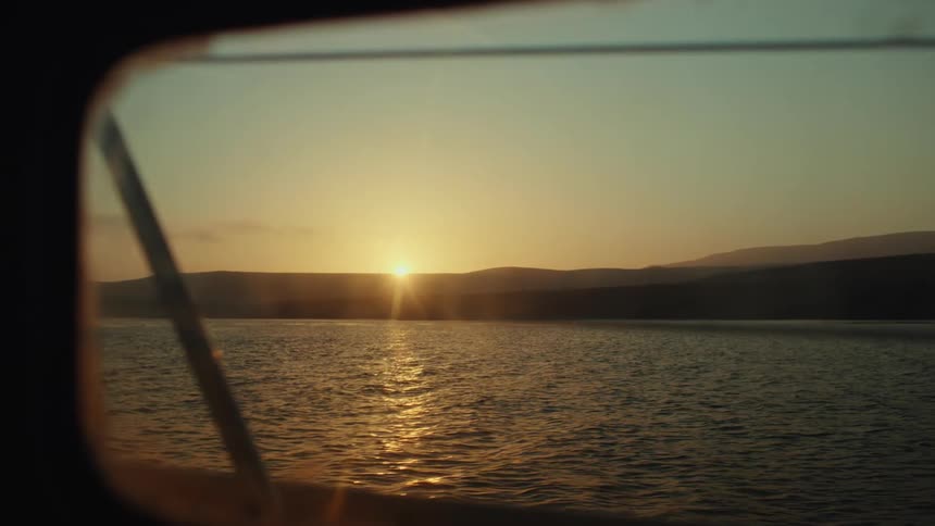 Sun shines from above the hills on the horizon and into the window of a passing boat