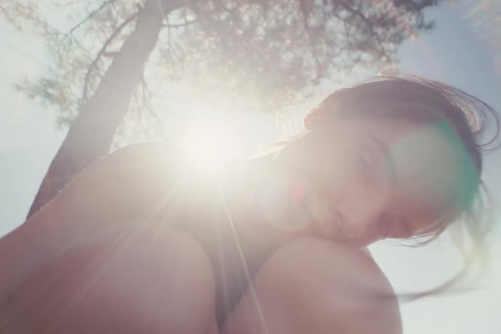 Young woman rests her arm on her knee looking forward under sunlight near a tree