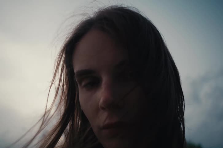 A woman with a serious expression her hair blowing in the wind against a cloudy sky
