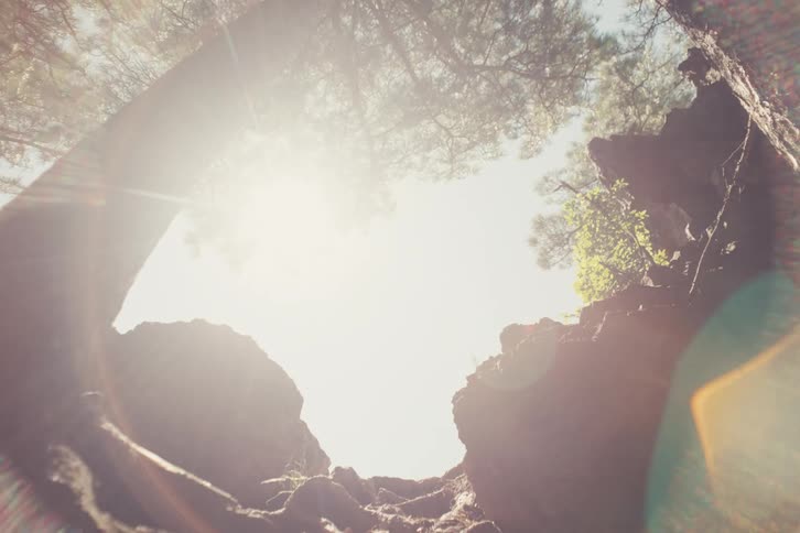 A person is seen in silhouette climbing upward through rocks and trees with bright sunlight streaming dramatically around them