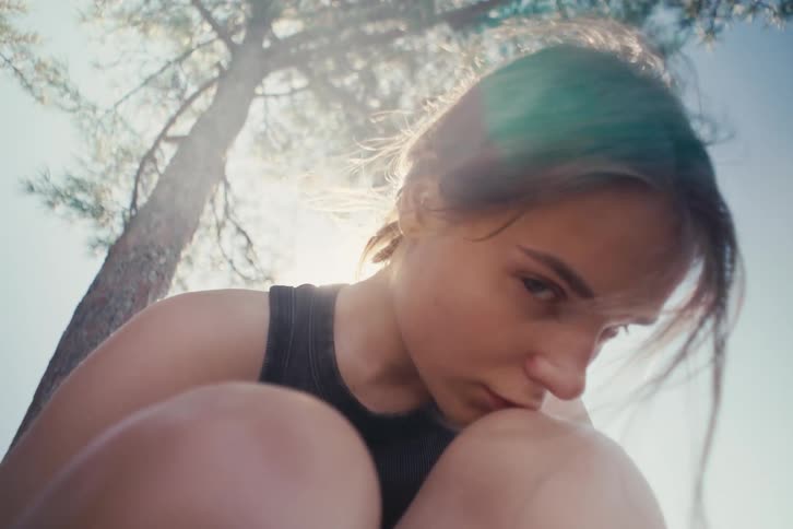 Young woman rests her face on her knee as sunlight filters through the tree leaves in the background