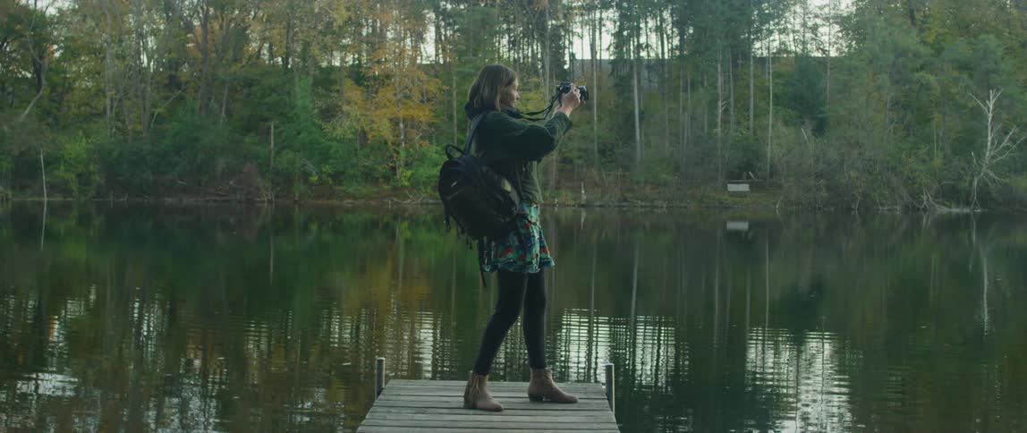 A woman photographs a lake and forest from a dock at sunset