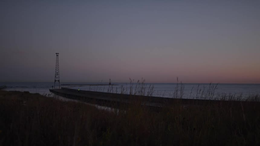 A tall metal tower stands at the edge of a pier extending into the calm sea at dusk with grasses in the foreground