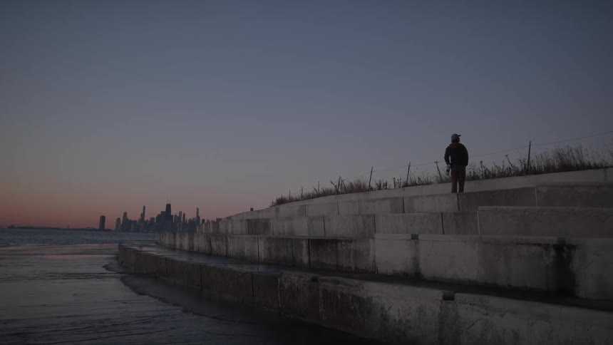 A man is standing by the shore at sunrise with chicago skyline in the background