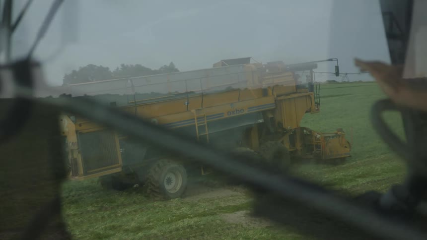 The video shows two large farming machines likely harvesters in a green field the machine in the foreground is partially obscured by the vehicle the camera is in and a person can be seen operating its controls the second machine which is yellow and