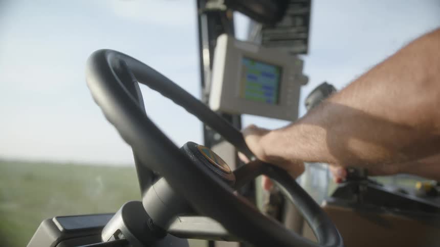 A persons hands operating the steering wheel and a joystick controller inside the cabin of a tractor