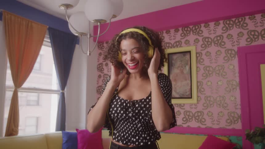 A video shows a young woman with curly brown hair dancing in a pink room she is wearing a black and white polkadot top black pants and yellow headphones she is smiling and appears to be enjoying herself