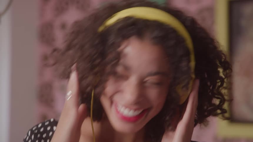 A joyful woman with curly hair smiles and dances happily while listening to music on her bright yellow headphones in her room