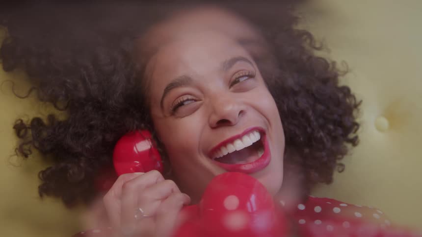 A young woman overly excited sits on a yellow chair holding a red vintage telephone