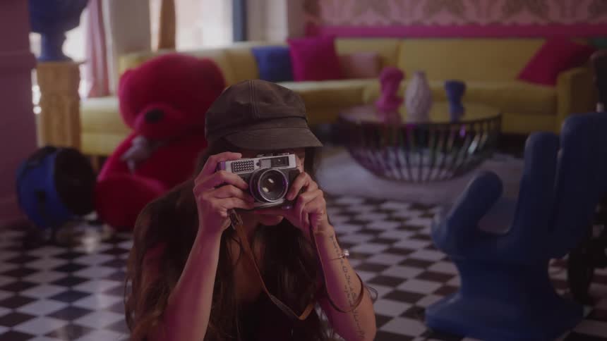A video shows a young woman with a long brown hair wearing a brown cap and a dark tank top she is holding a vintage camera and appears to be taking pictures in a room with a checkerboard floor