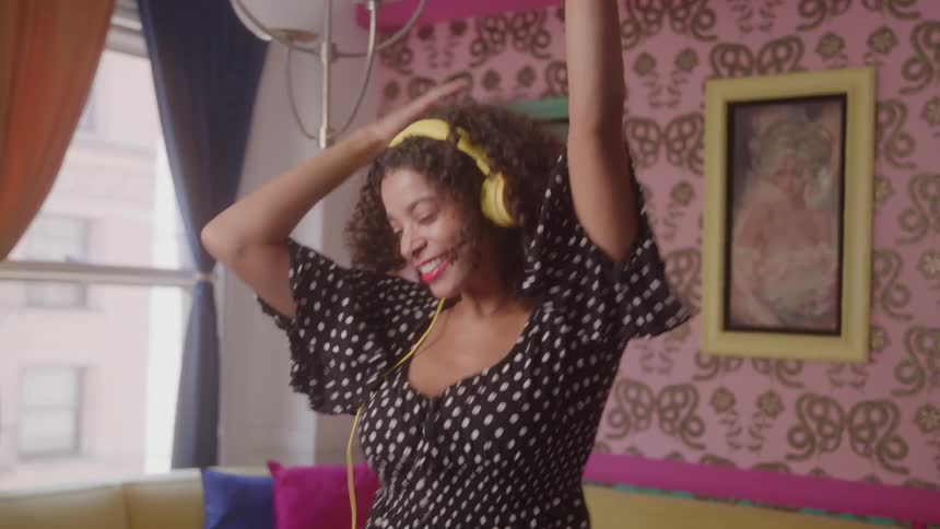 A video shows a young woman with curly brown hair wearing a black and white polkadot top and yellow headphones she is happily dancing in a room with pink patterned wallpaper smiling and moving her arms to the music