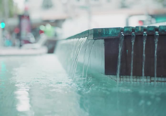 A video shows a closeup shot of a modern fountain with a soft outoffocus background the water cascades over the edge of the fountain creating a gentle flow and ripples in the pool below