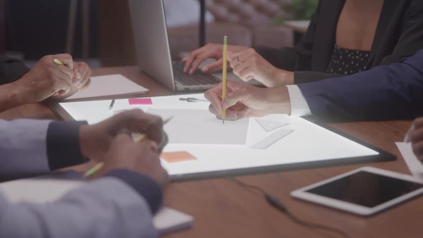 People sit around a table working collaboratively with one using a laptop and others writing with pencils on a brightly lit surface
