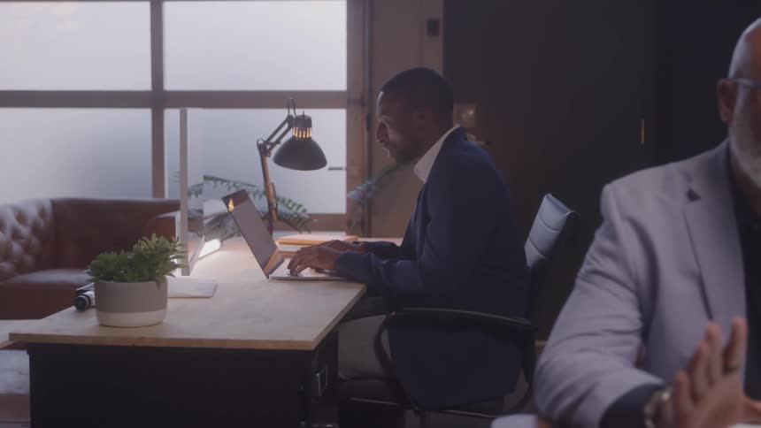 Man in a suit working on a laptop at a desk in an office with another man blurred in the foreground