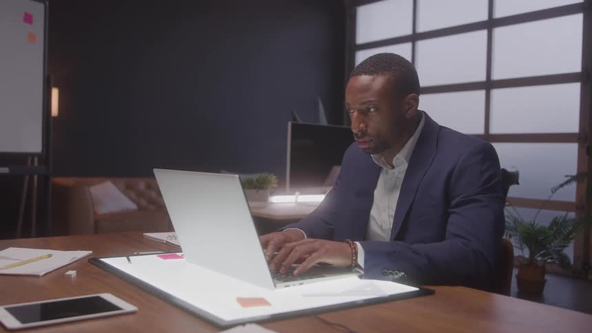 A welldressed professional man working late at a modern office desk typing intently on his laptop computer