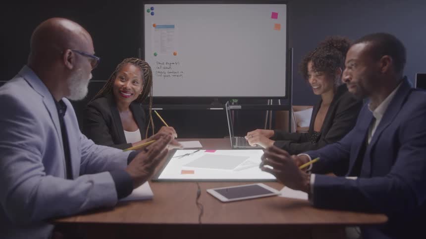 Group of four people in a meeting around a table with a whiteboard in the background