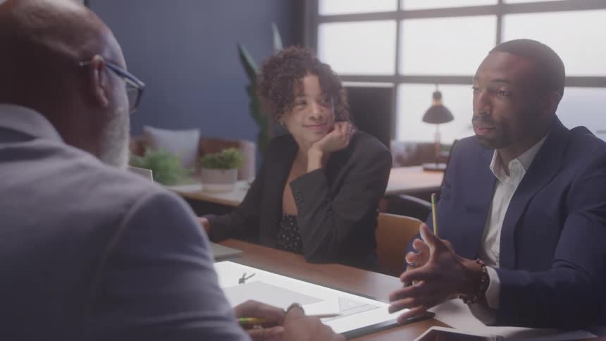 Three professionals sit at a table in discussion with one man gesturing as he speaks while a woman smiles attentively