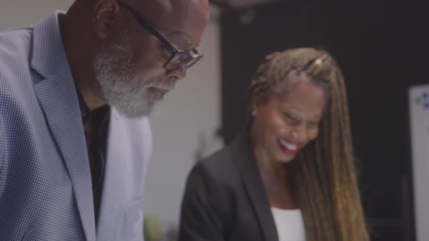 A man in glasses and a light blazer focuses on his work while a woman beside him smiles brightly