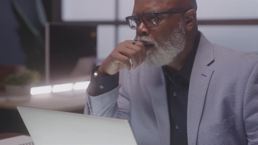 An older man in glasses and a light blazer sits at a desk thoughtfully resting his hand near his mouth while looking at a laptop screen