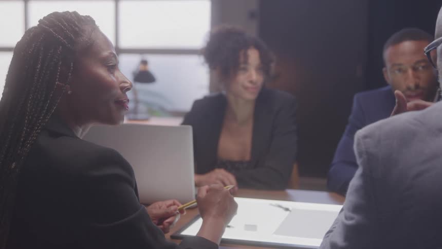 Three professionals in business attire sit around a lit workspace engaged in conversation