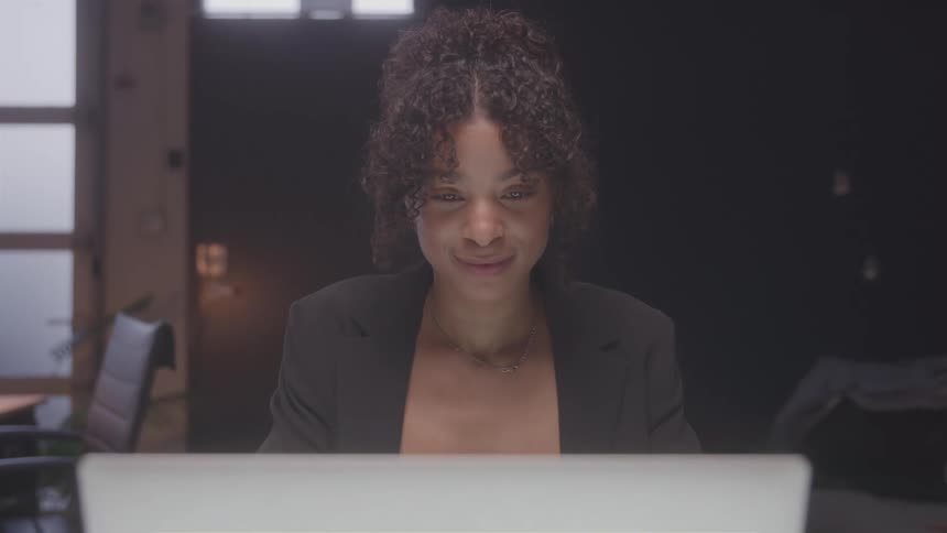 A young woman with curly hair wearing a blazer suit sits in an office typing on a laptop