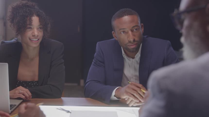 A woman and a man in business attire sit at a table engaged in discussion with an older man across from them