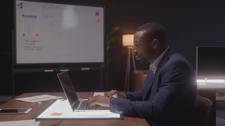 A welldressed man working late at night in a modern office typing on a laptop with a whiteboard and papers visible in the background