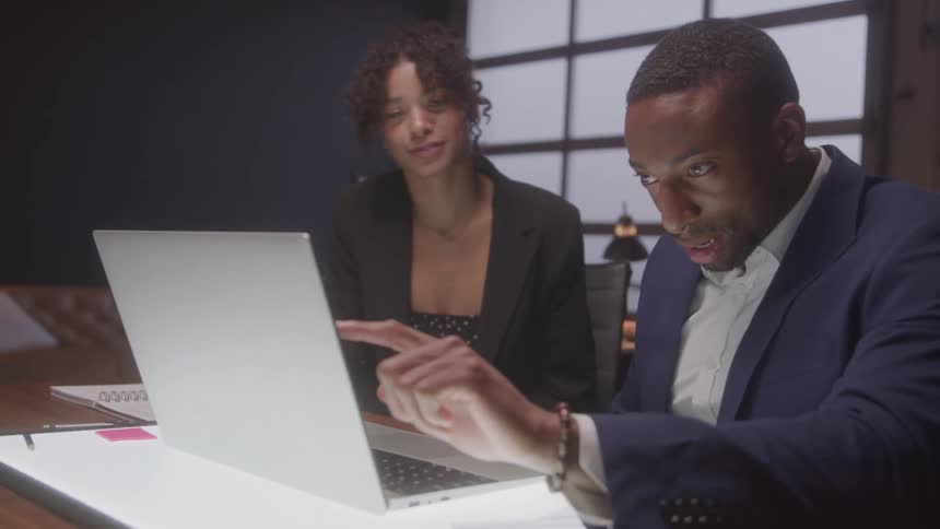 A young woman and a young man sit behind a desk in a conference room both wearing blazer suits as the woman explains something on a laptop screen in front of them