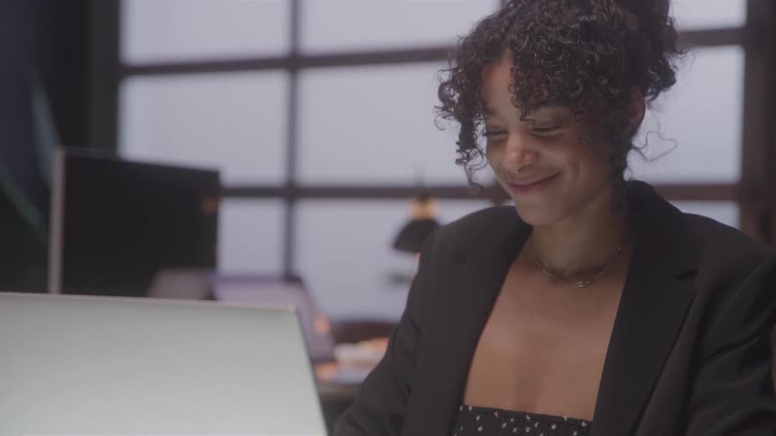 A professional woman with curly hair wearing a blazer is seen from the chest up smiling and looking down at a laptop screen in a dimly lit office setting